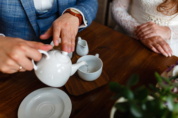 Man in a fashionable suit pours tea from a teapot in a mug. Rules of etiquette in a cafe. Man takes care of a woman. A young couple in the loft cafe drinking coffee and tea sitting at a wooden table.