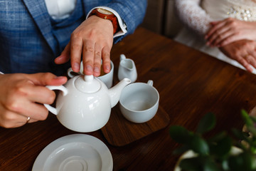 Man in a fashionable suit pours tea from a teapot in a mug. Rules of etiquette in a cafe. Man takes care of a woman. A young couple in the loft cafe drinking coffee and tea sitting at a wooden table.