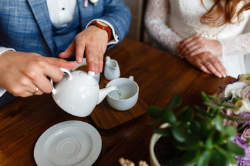 Man in a fashionable suit pours tea from a teapot in a mug. Rules of etiquette in a cafe. Man takes care of a woman. A young couple in the loft cafe drinking coffee and tea sitting at a wooden table.