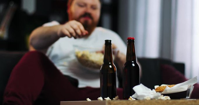 Smiling fat man with beard watches TV in the room and eats pop-corn at the table with beer. Fat man drinks beer. Concept: Malnutrition, sedentary lifestyle, rest on the couch, obesity