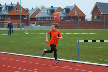 Obraz premium Little boy in orange outfit runs on the running track while he looks back at his competitors.