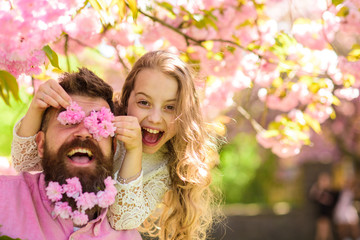 Fototapeta premium Dziecko i mężczyzna z delikatnymi różowymi kwiatami w brodzie. Dziewczyna z tatą w pobliżu kwiatów sakura na wiosenny dzień. Ojciec i córka na szczęśliwej twarzy bawią się kwiatami jako okulary, tło sakura. Pojęcie czasu dla rodziny
