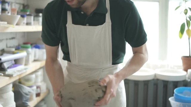 Male ceramist is preparing white loam in bright studio. Professioanl is holding the block of clay in his hands and kneading it by his fingers. Man in dickey is standing near shelves with sculptures