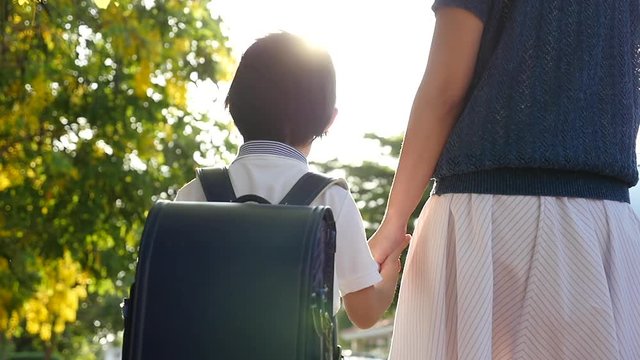 Asian mother holding hand of little son with backpack outdoors, back to school slow motion 