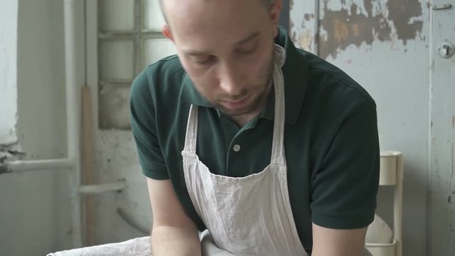 Portrait of ceramist, who is working on throwing wheel in his studio. Man is sitting at the equipment with a piece of white clay on it and forming it up and down by his hands, additing water. Male