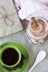 Oatmeal, flavored with yogurt, sprinkled with cinnamon in a glass jar. Next is a cup of coffee. Next is a craft notebook and a pink napkin.