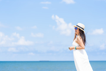 Left side of women in white dress and hat walking along the beach with blue sea and sky front view. Travel and summer conceptual.
