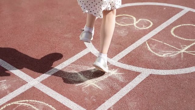 Tracking shot of a girl jumping while playing Hopscotch at park. Little cute girls on children playground outdoors. Legs of kids jumps hopscotch on asphalt. 