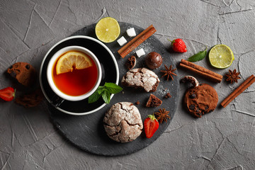 Fragrant tea in a black cup on a black plate with biscuits, lemon, cinnamon and fruits.