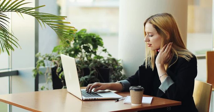 Young blonde girl works with a laptop sittig at the table in cafe. Freelancer is looking for a job, printing something and drinking coffee