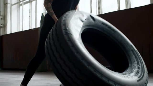 Tracking shot of determined young woman with tattoos pushing and flipping heavy tire in empty gymnasium