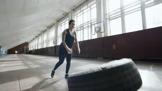 Tracking of strong sportswoman breathing hard and pushing and flipping heavy tire in empty gymnasium