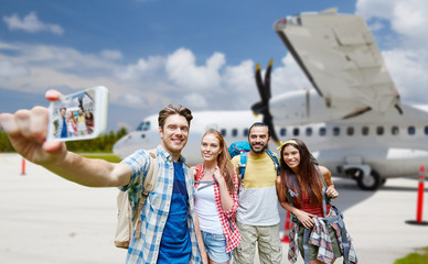air travel, tourism and technology concept - group of smiling friends with backpacks taking selfie by smartphone over plane on airfield background