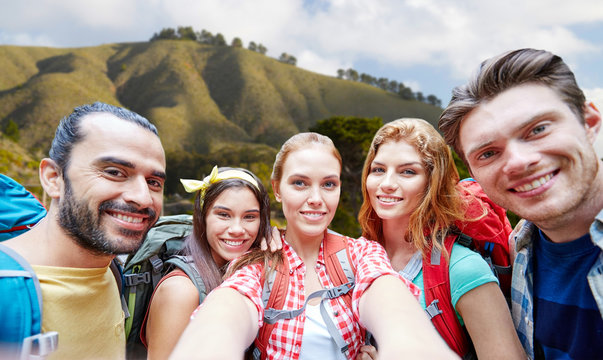 technology, travel, tourism, hike and people concept - group of smiling friends with backpacks taking selfie over big sur of california hills background