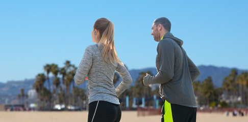 fitness, sport and technology concept - happy couple running and listening to music in earphones over venice beach background in california
