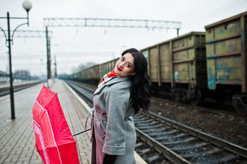 Fototapeta premium Brunette girl in gray coat with red umbrella in railway station.