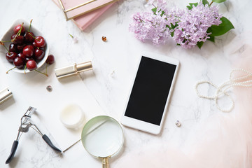Overhead image of feminine workspace mock up with phone screen, accessories and lilac flowers arranged on marble background