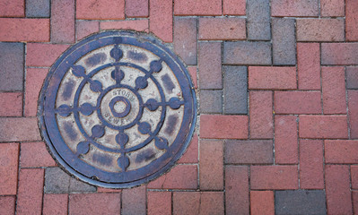 Close up photo of a storm sewer manhole cover on a brick sidewalk.