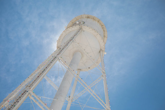 Looking up at a bright white water tower from below backlit with the sun and a bright blue sky.