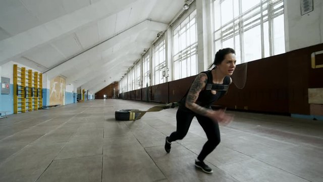 Tracking of determined sportswoman with tattoos running and pulling heavy tire while cross-training in empty gymnasium