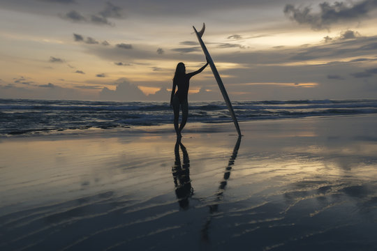 Full length of silhouette young woman with surfboard standing at beach against sky during sunset