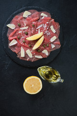 Stone slate tray with carpaccio made of raw marbled beef, above view over black stone background,...