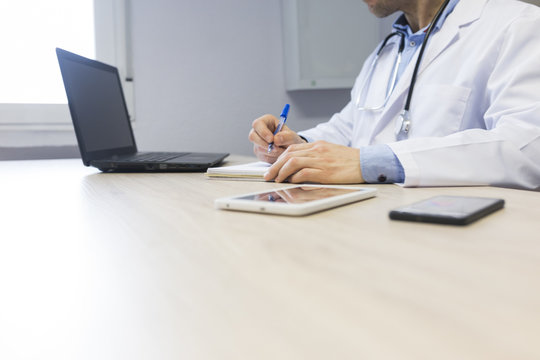 Young Doctor At The Office Writing On Notebook. Laptop, Tablet And Mobile Phone On The Table. Medical Concept