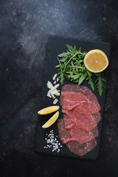 Carpaccio made of marbled beef served with rucola, lemon and parmesan on a stone slate. Above view on a dark scratched metal background, vertical shot