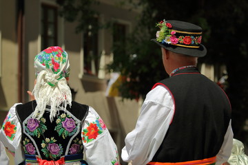 Kobieta i mężczyzna (górne części ciała), z tyłu, stoją, ubrani w odświętne łowickie stroje, pan w ozdobionym kwiatami kapeluszu, kobieta w wyszywanej w kwiaty chustce na głowie, czarne serdaki - obrazy, fototapety, plakaty