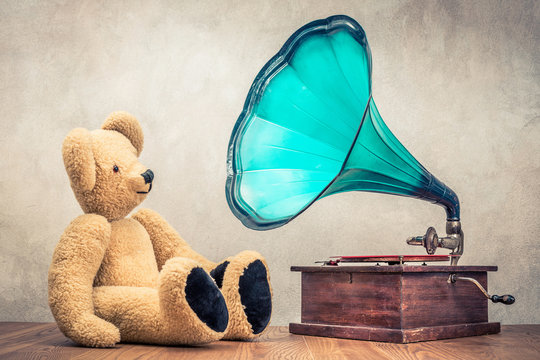 Retro Teddy Bear toy sitting near classic old gramophone turntable player on oak wooden floor front concrete wall background. Listening to music concept. Vintage instagram style filtered photo