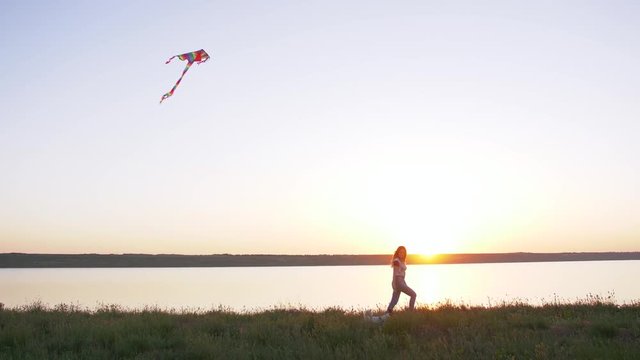 Young happy woman and het little dog walking with flying kite on a glade at sunset, slow motion