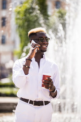 Portrait of smiling young afro american man talking on mobile phone and drinking phone while standing near the fountain in the city