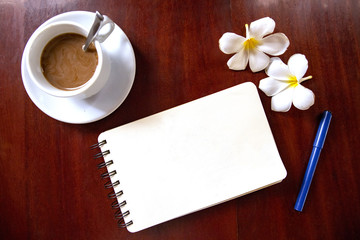Obraz na płótnie Canvas Blank spiral notebook page and pen, coffee cup and frangipani flowers top view photo on wooden table.