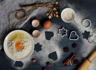 A set for making cookies. Flour and egg in a bowl, butter, sugar, cookie shapes, nutmeg, cinnamon, anise, rolling pin, whisk for whipping. The dark background is peppered with flour. Close-up.