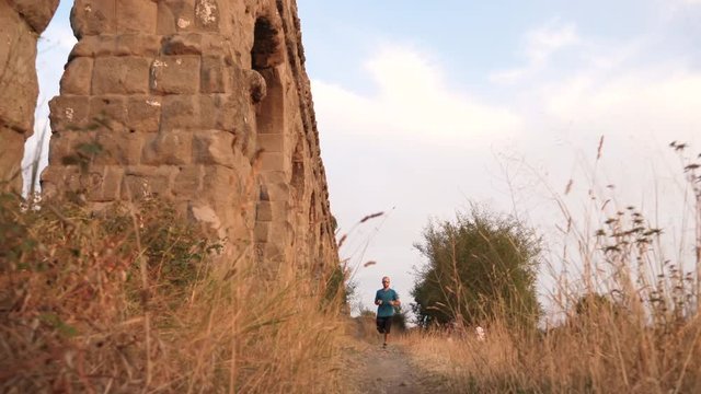 Young man in sportswear running along ancient roman aqueduct Appio Claudio. Training outdoor alone in peaceful park in Rome at sunset slow motion.