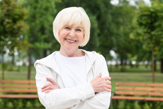 smiling senior woman with crossed arms standing in park