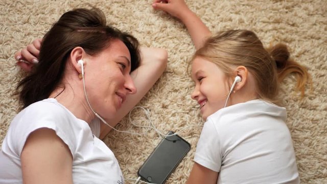 Funny footage of young woman with little daughter lying on the floor looking at each other and listening to music on the phone, they use one earphone.