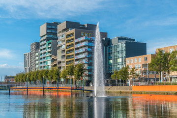 Fototapeta na wymiar Lelystad, October 27th 2017: locals walking and crossing the pond with fountain in the center with modern business buildings and houses in the background