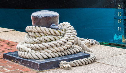 iron bollard with heavy mooring lines on a red brick walkway next to a blue and black ship's hull...