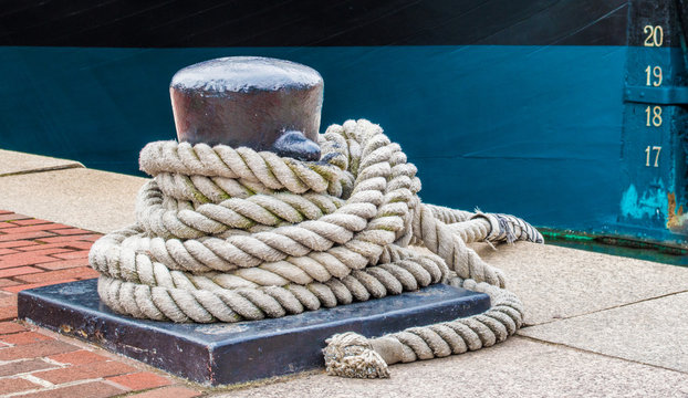iron bollard with heavy mooring lines on a red brick walkway next to a blue and black ship's hull with draft marks