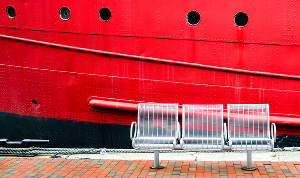 3 empty white metal seats on a red brick walkway next to the red hull of a large vintage ship with copy space