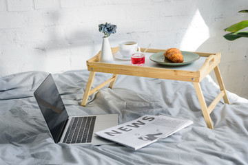 laptop with blank screen and breakfast with croissant and coffee on tray on bed in morning