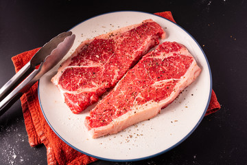 Two cuts of strip steak resting on a plate being prepared with salt and pepper for cooking.