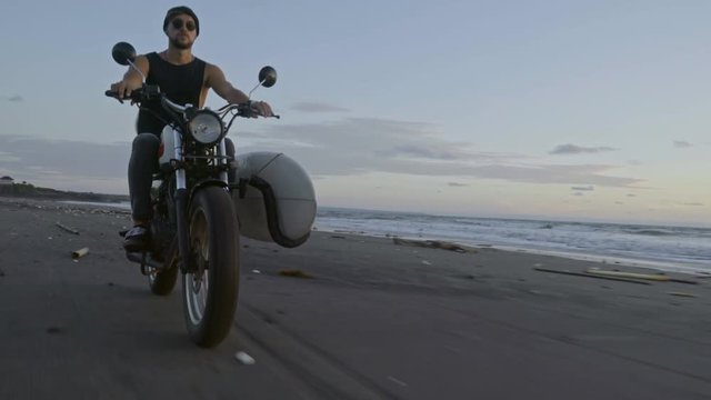 Front view of young man in beanie and sunglasses riding motorbike with surfboard attached to it during sunset at the beach