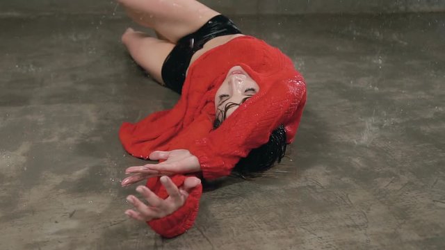 Wet young woman in red sweater and black shorts is dancing on the floor under the rain and splashes of water. Drops water, rain, contemporary dance