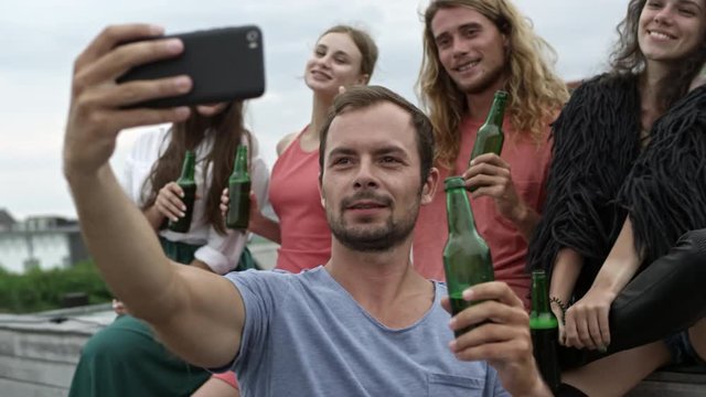 Young friends holding bottles of beer and taking selfie together, posing and waving for smartphone camera