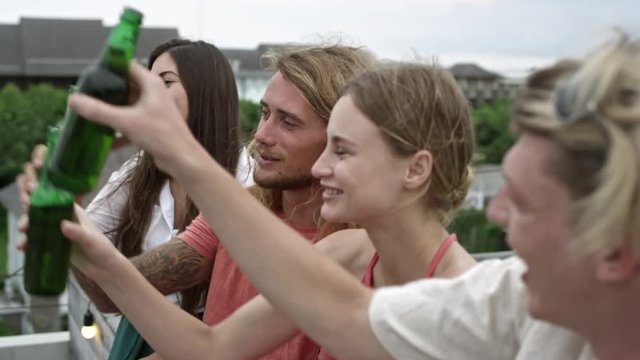 Group of young people spending time outdoors, clinking glass bottles of beer, drinking and smiling