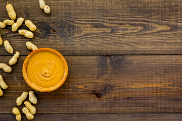 Product for hearty breakfast. High-calorie product. Peanut butter in bowl near nuts in shell on dark wooden background top view space for text