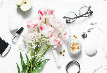 Flat lay women's accessories set - tulips bouquet, cosmetics, glasses, phone, apple, headphones on light background top view. Beauty and fashion concept