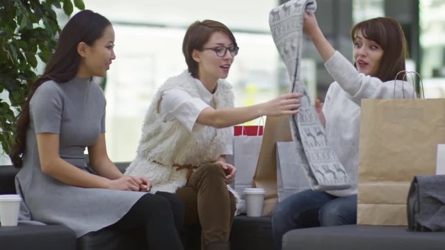 Medium shot of three young women sitting on sofa in shopping mall, one of them showing new scarf with reindeer pattern to her friends